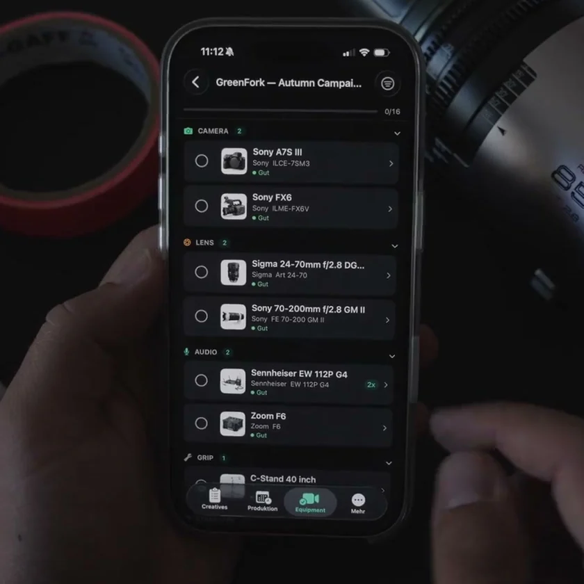 The TillyBoard iPhone app with the packing list GreenFork Autumn Campaign, grouped into camera, lens, audio and grip, 0 of 16 packed. Next to it a roll of gaffer tape and an 85 mm cine lens.